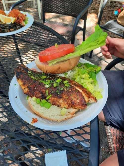 A seasoned fish fillet on a bun with lettuce and tomato, served on a white plate on a table.