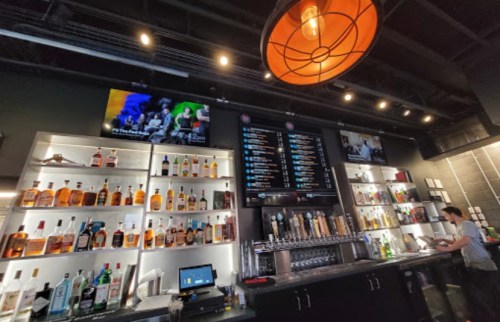 A modern bar with shelves of liquor, a large menu board, and a bartender preparing drinks under warm lighting.