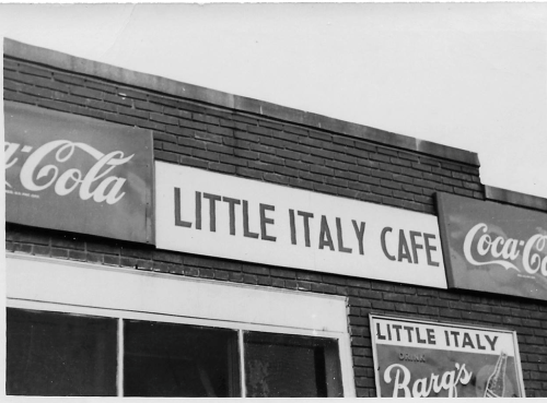 Signage for "Little Italy Cafe" with Coca-Cola advertisements on a brick building. Black and white photo.
