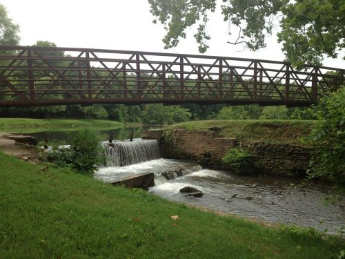 A metal bridge spans a small waterfall, surrounded by lush greenery and a calm river.