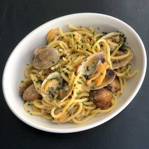 A bowl of spaghetti with clams, garnished with parsley on a dark surface.