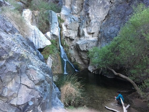A serene waterfall cascades into a clear pool surrounded by rocky cliffs and lush greenery, with a person sitting nearby.