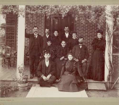 A historical black-and-white photo of a large group of people posed on a porch, dressed in 19th-century clothing.