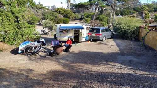 A camper and minivan parked in a gravel area, with a person sitting nearby and bicycles leaning against a tree.