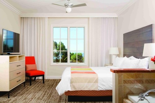 A cozy hotel room featuring a bed, TV, chair, and large window with a view, decorated in neutral tones.