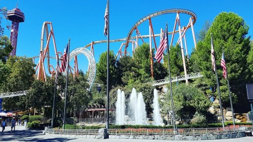 A vibrant amusement park scene featuring roller coasters, fountains, and flags under a clear blue sky.