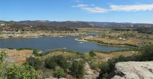 A scenic view of a lake surrounded by hills and greenery under a blue sky with scattered clouds.