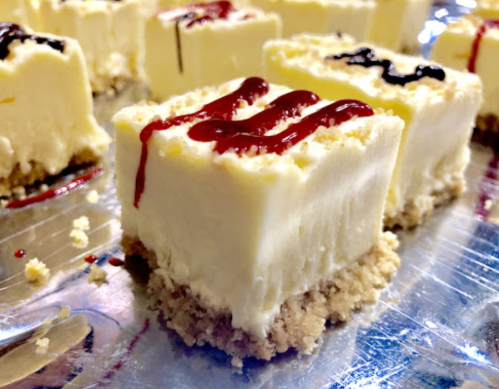 Close-up of creamy cheesecake squares with a graham cracker crust, drizzled with red and black sauces.