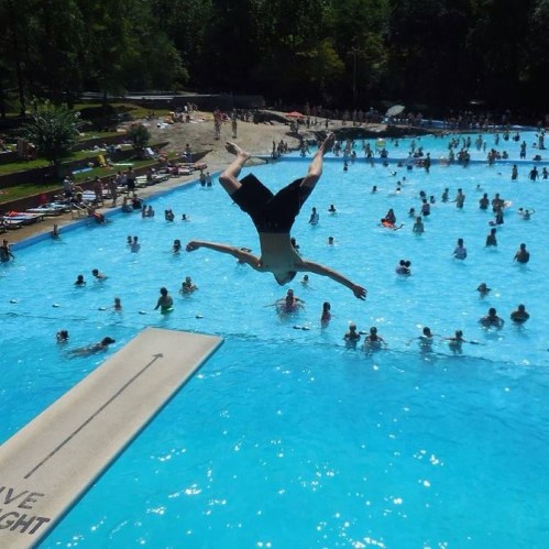 A person jumps off a diving board into a crowded pool on a sunny day, surrounded by swimmers and greenery.