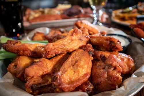 A close-up of crispy chicken wings served with celery and dipping sauce, with drinks and other food in the background.