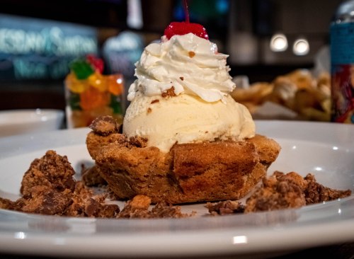 A dessert featuring a cookie bowl filled with ice cream, topped with whipped cream and a cherry, surrounded by crumbled cookies.