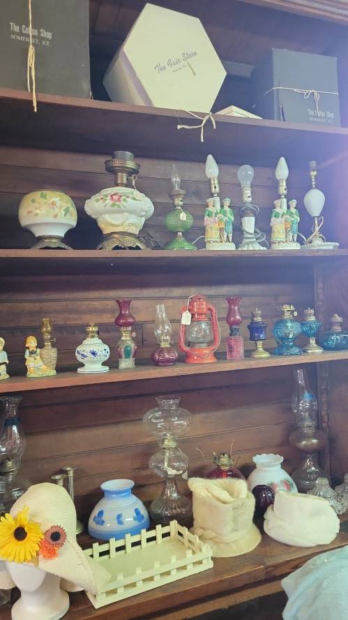 A wooden shelf displaying various vintage lamps, decorative items, and hats in a quaint shop setting.