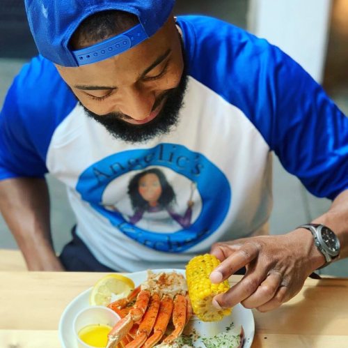 A man in a blue cap enjoys a plate of crab legs, corn, and lemon, smiling as he prepares to eat.