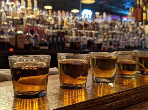 A row of shot glasses filled with dark liquid on a wooden bar, with a blurred background of liquor bottles.