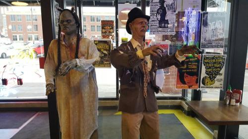 Two life-sized zombie figures in a shop window, one in tattered clothing and the other in a suit, surrounded by posters.