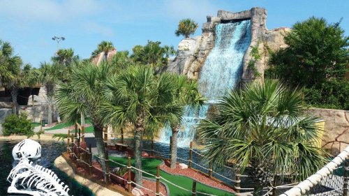 A mini-golf course featuring a waterfall, palm trees, and lush greenery under a clear blue sky.
