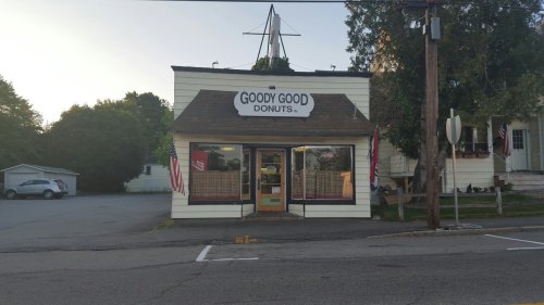 A quaint donut shop named "Goody Good Donuts" with a sign and flags, set in a peaceful neighborhood.
