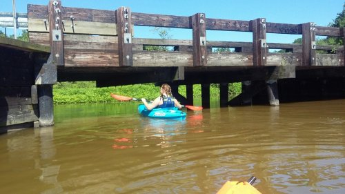 A person in a blue kayak paddles under a wooden bridge on a calm, brown river.