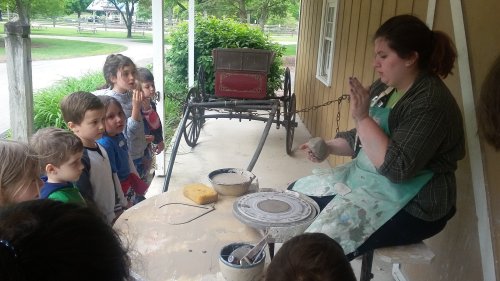 A pottery instructor demonstrates techniques to a group of attentive children at an outdoor workshop.