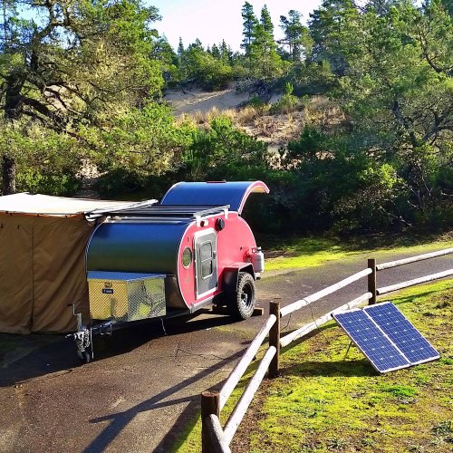 A small, red and silver camper trailer parked near a wooden fence, with solar panels nearby and trees in the background.