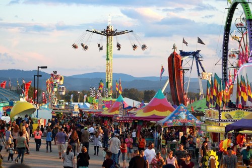 A lively fair scene with colorful rides, tents, and crowds enjoying the festivities under a sunset sky.