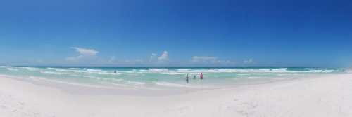 A panoramic view of a sunny beach with gentle waves and people enjoying the water under a clear blue sky.