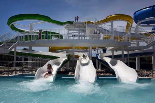 Three water slides at a water park, with children splashing into a pool below and colorful slides in the background.