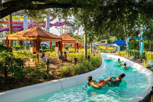 People relax on inner tubes in a lazy river at a water park, surrounded by greenery and colorful slides in the background.