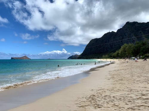 A scenic beach with golden sand, clear blue water, and distant mountains under a partly cloudy sky. People enjoy the shoreline.