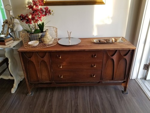 A wooden sideboard with three drawers, decorative items, and a plant on top, set against a light wall.