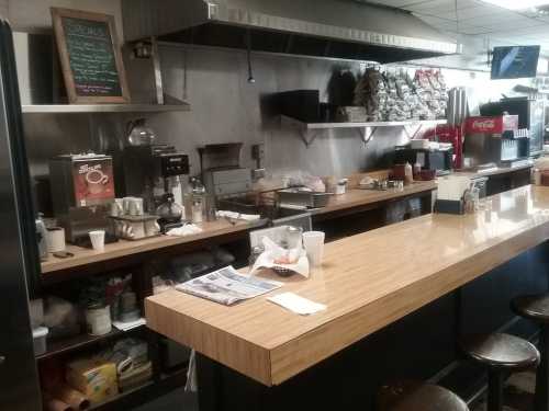 A diner counter with a wooden surface, coffee machines, and a chalkboard menu, featuring stools and scattered items.