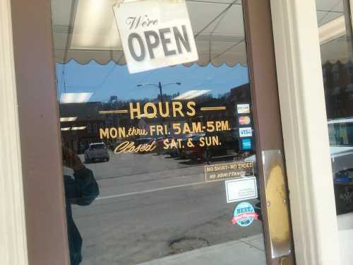 A storefront door with an "Open" sign and business hours: Mon-Fri 5 AM-5 PM, closed Sat & Sun.