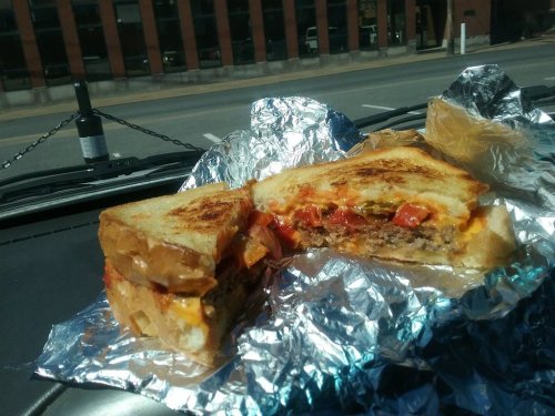 A grilled sandwich with layers of meat, cheese, and vegetables, wrapped in foil, resting on a car dashboard.
