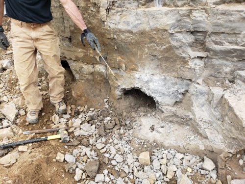 A person in work gloves stands near a rock face with a visible hole, surrounded by loose stones and tools.