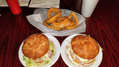 Two burgers with lettuce on white plates, accompanied by a serving of onion rings and a drink in the background.