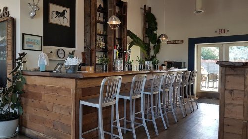 A cozy bar area with wooden stools, shelves of drinks, and large windows letting in natural light.