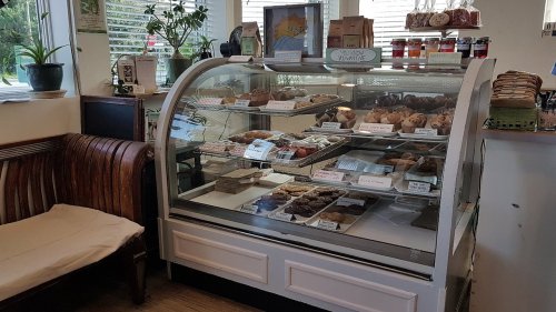 A display case filled with various baked goods, with a cozy seating area nearby and plants in the background.