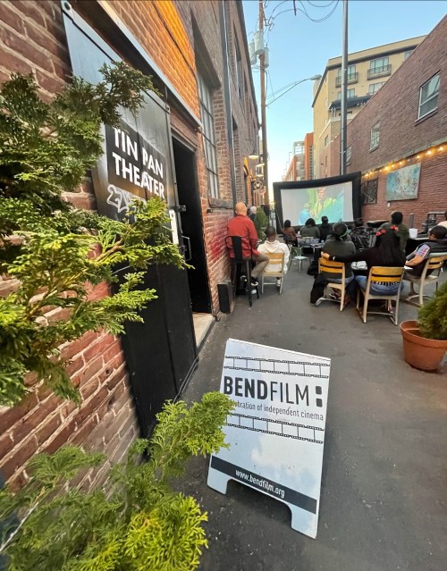 Outdoor screening at Tin Pan Theater, with an audience watching a film in an alley, surrounded by buildings and greenery.