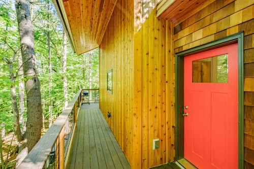 A wooden exterior of a house with a bright red door, surrounded by trees and a wooden walkway.