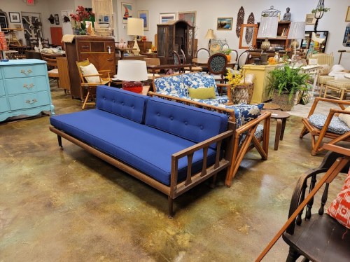A blue upholstered sofa with wooden frame in a vintage furniture store filled with various decor items.