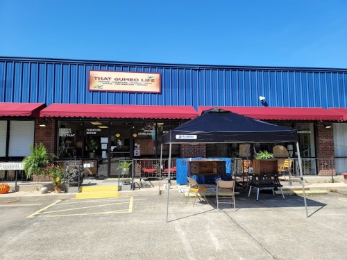 A blue storefront with a sign reading "That Gumbo Life" and a tent set up outside, featuring tables and chairs.
