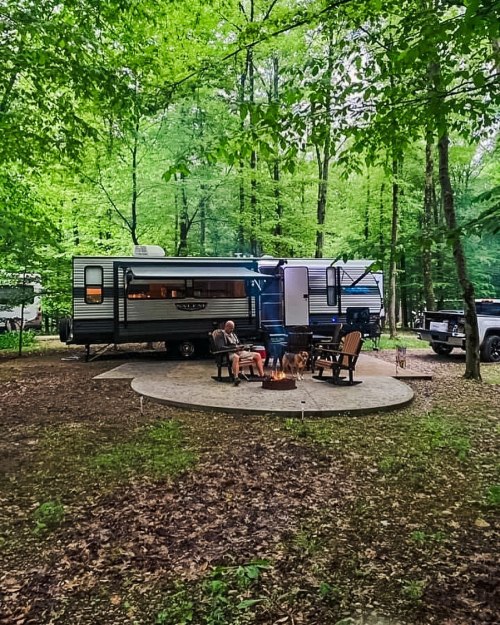A camper parked in a wooded area, with two people and a dog sitting around a fire pit on a circular patio.