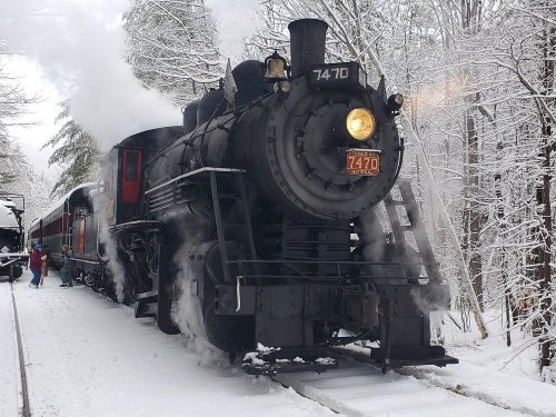 A vintage steam train with smoke billowing, surrounded by snow-covered trees in a winter landscape.