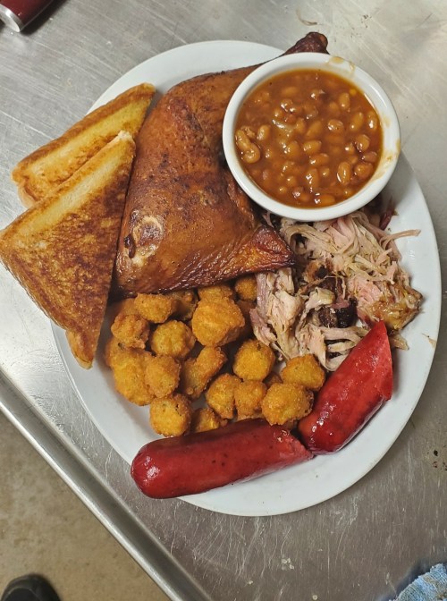 A plate of food featuring roasted chicken, pulled pork, baked beans, cornbread, fried okra, and sausage.