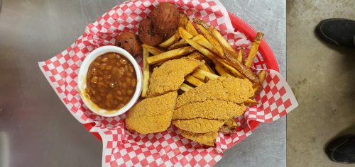 A basket of fried fish, French fries, baked beans, and cornbread muffins on a checkered red and white paper.