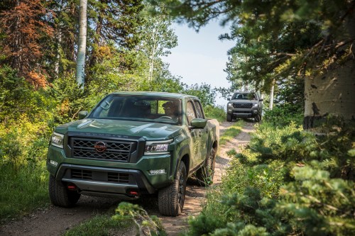 Two trucks driving on a narrow dirt path through a lush green forest.
