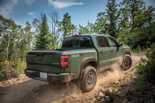 A green pickup truck driving off-road on a dusty trail surrounded by trees.