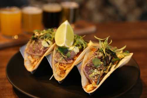 Three tacos filled with meat, topped with herbs and a lime wedge, served on a black plate with drinks in the background.