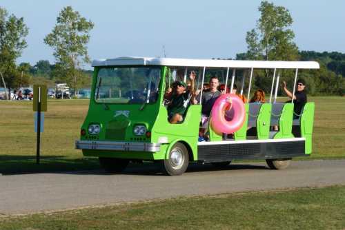 A green open-air shuttle bus with passengers, some holding a large pink inner tube, driving through a grassy area.