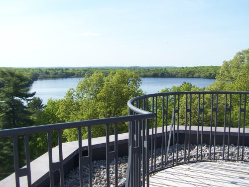 View from a balcony overlooking a serene lake surrounded by lush green trees under a clear blue sky.